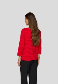 Frau mit langen braunen Haaren, die ein rotes, strukturiertes Langarmoberteil und schwarze Hosen trägt, steht mit dem Rücken zu einem grauen Hintergrund.