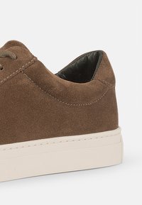 Zapatillas de ante marrón con un diseño minimalista, que presentan una suela de goma blanca a contraste y un interior negro. Cordones y detalles de costura visibles.