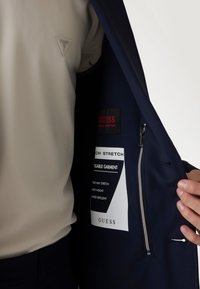 Tengerészkék dzseki cipzáras zsebből, fehér címkével, amely gondozási utasításokat tartalmaz; alatta világos bézs ing, texturált anyagból.