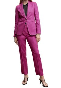 Blazer rose cintré et pantalon assorti en tissu lisse, avec une fermeture à un bouton et une coupe droite. Haut noir en dessous.