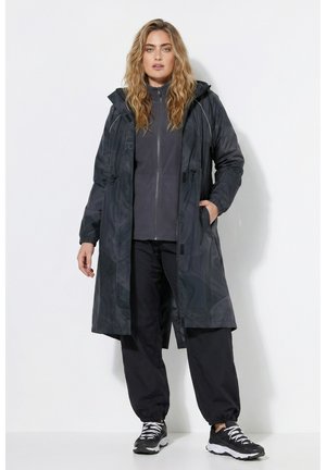 Woman with wavy blonde hair wearing a long black hooded coat, dark zip-up jacket, loose black pants, and black sneakers with white soles.