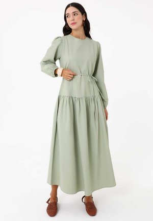 Femme portant une longue robe vert sauge à manches bouffantes et une ceinture à la taille, associée à des ballerines tressées marron et des boucles d’oreilles créoles.