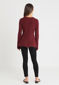 Donna con lunghi capelli castani, che indossa un maglione attillato bordeaux con orlo scallop, pantaloni skinny neri e sandali con tacco aperti neri, in piedi con la schiena rivolta.
