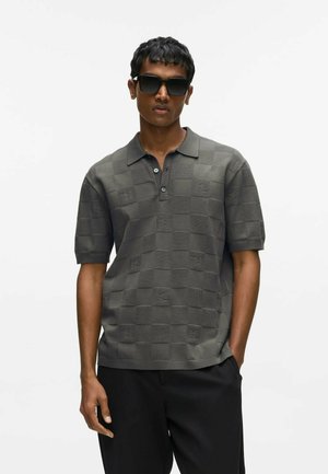 Homme portant des lunettes de soleil foncées, un polo à manches courtes à carreaux gris, et un pantalon noir, debout avec une main dans la poche.