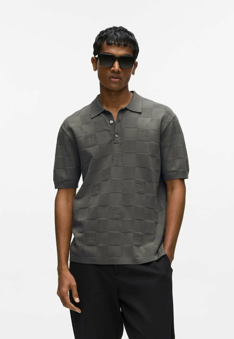 Homme portant des lunettes de soleil foncées, un polo à manches courtes à carreaux gris, et un pantalon noir, debout avec une main dans la poche.