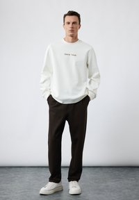 Hvid sweatshirt med teksten "Liberté Totale", parret med mørkebrune bukser og hvide sneakers, sat mod en neutral baggrund.