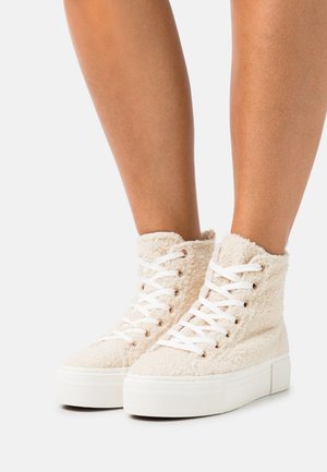 Witte hoge sneakers met dikke plateauzolen en een zachte, gestructureerde stof, gedragen op blote benen tegen een witte achtergrond.