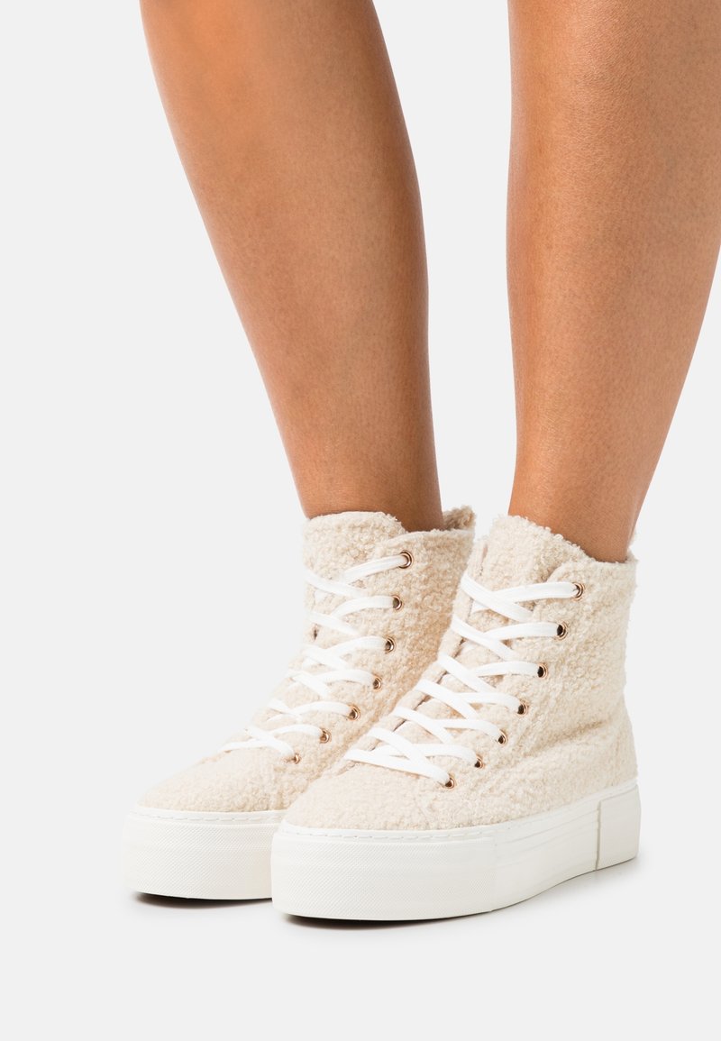 Baskets montantes blanches avec semelles épaisses compensées et tissu texturé moelleux, portées sur des jambes nues sur un fond blanc.