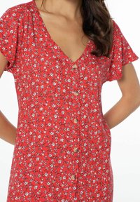Robe rouge à fleurs avec manches courtes volantées, encolure en V et fermeture à boutons sur le devant. Le motif floral présente de petits accents blancs et noirs.