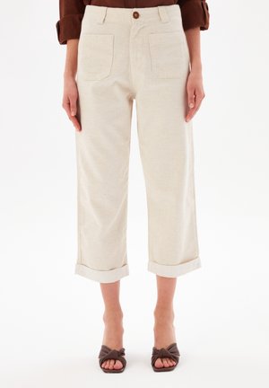 Vrouw draagt crèmekleurige cropped wijde broek met voorzakken en bruine open-teen haksandalen op een witte achtergrond.