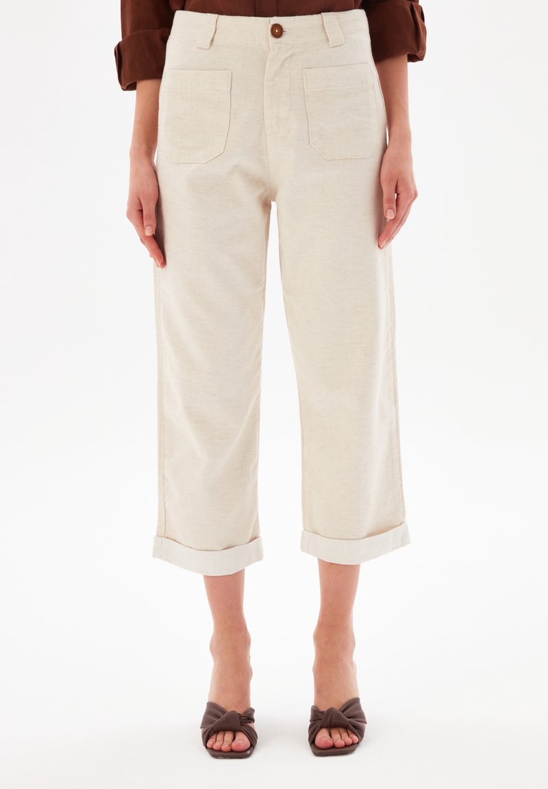 Femme portant un pantalon crème court à jambes larges avec poches plaquées devant et sandales à talons ouvertes marron sur fond blanc.