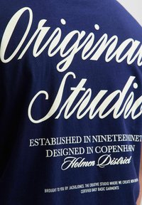 Tamnoplava pamučna majica s bijelim tiskanim tekstom u kurzivnom i standardnom fontu, detaljizirana s informacijama o marki i lokaciji na stražnjem dijelu.