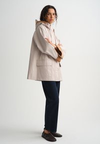 Žena stoji prekriženih ruku, nosi bež jaknu s kapuljačom, tamne hlače i tamne slip-on cipele, na jednostavnoj svijetloj pozadini.