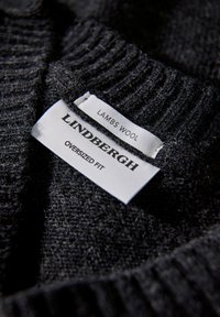 Svart lammullströja med en strukturerad yta. Etiketterna visar "LINDBERGH", "LAMMSPULLOVER" och "OVERSIZED FIT" i vitt på kontrasterande tyg.