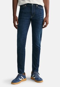 Jeans in denim blu scuro con una vestibilità slim, caratterizzati da cuciture arancioni e un classico design a cinque tasche. Indossati con sneakers blu e dettagli bianchi.