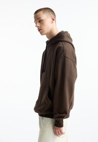 Brauner Pullover-Hoodie mit einer Vordertasche, Kordelzugkapuze und gerippten Bündchen. Glatte Textur und entspannte Passform, präsentiert an einem Modell.