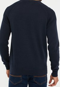 Marineblauwe gebreide trui met geribbelde schouderaccenten en boorden, ronde halslijn en een gestructureerde afwerking. De achterzijde toont minimale details.