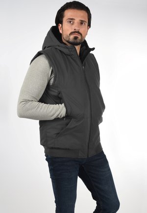 Svart ermeløs vest med hette, med glidelås foran og sidelommer. Brukt over en lys grå langermet skjorte og kombinert med mørke jeans.