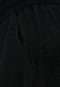 Közelkép fekete, texturált anyagú nadrágról rugalmas derékrésszel és látható oldalzsebbel.