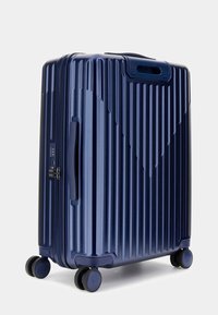 Dunkelblauer Hartschalenkoffer mit vertikalen Rillen, Seitengriff, Schlösserfeld und vier Rollen für Mobilität. Glatte Textur und elegantes Design.
