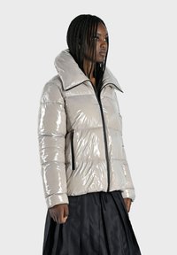 Doudoune matelassée en beige clair brillant avec un col montant, des détails de fermeture éclair noirs et des poches latérales ; la texture matelassée renforce la chaleur.