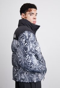 Jakna s punjenjem u crnoj i bijeloj boji s uzorkom; s visokim ovratnikom i zatvaranjem na patentni zatvarač. Materijal izgleda glatko s kutijama od quiltera.