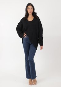 Suéter negro de gran tamaño con escote redondeado y puños acanalados, combinado con jeans oscuros de campana y sandalias de tacón transparente.