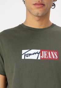 Olivno zelena bombažna majica ima pravokotni grafični na rdeči in beli barvi z besedilom "Tommy Jeans" in stiliziranim logotipom v modri barvi.