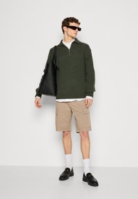 Maglione verde a maglia con texture e mezza cerniera, abbinato a pantaloni cargo beige. Una borsa a tracolla nera e scarpe, calzini bianchi e occhiali da sole completano il look.