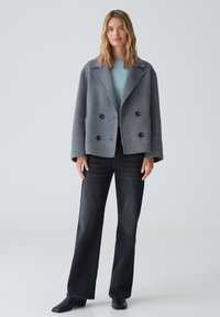 Grauer doppelt knöpfbarer Wollmantel mit großen Knöpfen, mit Reverskragen, über einem hellblauen Hemd, kombiniert mit schwarzen Jeans und schwarzen Stiefeletten.