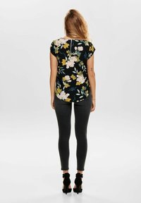 Blouse à imprimé floral avec manches courtes, fond noir et un ourlet arrondi, portée avec un jean noir slim et des chaussures à talons noires.