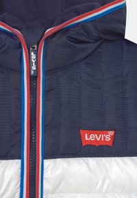 Námořnická modrá péřová bunda s bílou spodní částí, zipem s červenými a modrými pruhy a červeným logem Levi's na lemu. Texturovaný materiál.