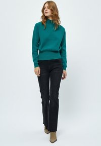 Maglione lavorato a maglia color teal con collo alto, abbinato a jeans neri a zampa d'elefante e stivali marroni, creando un outfit casual.