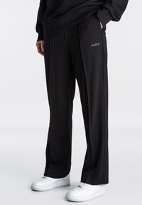 Schwarze Hosen aus glattem Stoff mit einer lockeren Passform, ausgestattet mit seitlichen Taschen und einem dezenten Logo-Detail auf der Vorderseite. Kombiniert mit weißen Sneakern.