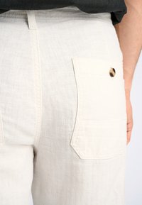 Witte linnen broek met een gestructureerde afwerking, voorzien van een achterzak die is beveiligd met een ronde knoop. Zichtbare stiksels langs de naden.