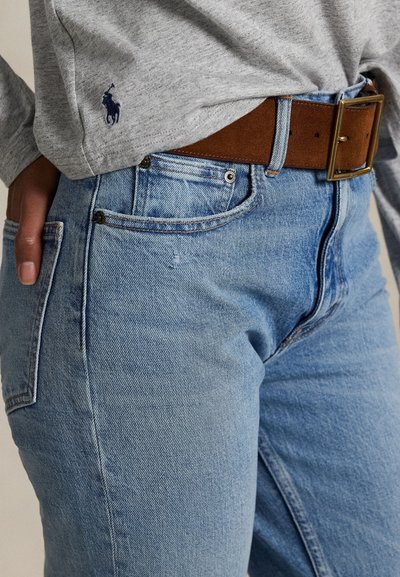 Személy világoskék farmert visel barna bőrvéssel és részben betűrt szürke hosszú ujjú inget.