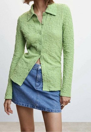 Femme portant une chemise boutonnée vert clair texturée et une mini-jupe en jean bleue, debout avec les mains détendues le long du corps.