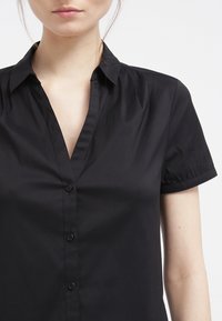 Camicia nera con colletto a V, maniche corte a palloncino e una texture liscia. Presenta un colletto classico e cinque bottoni abbinati.