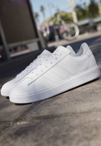Par hvide Adidas lave sneakers med snørebånd på en betonoverflade udendørs i dagslys.