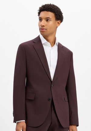 Mann trägt dunkelbraunen Anzugjacke und weißes Hemd, schaut seitlich vor schlichtem weißen Hintergrund.