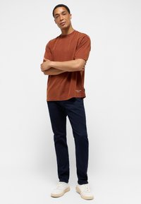 Koszulka z krótkim rękawem w kolorze rdzy z okrągłym dekoltem, zestawiona z ciemnoniebieskimi dżinsami i białymi trampkami.