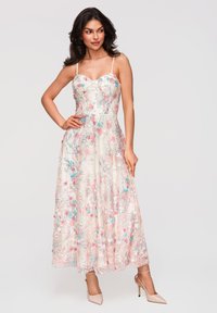 Femme portant une robe blanche sans manches, longueur cheville, avec une broderie florale rose et bleue, et des chaussures à talons hauts couleur chair, posant avec une main sur la hanche.