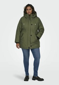 Olivgrön vadderad parka-jacka med huva, två framfickor, knappstängningar och dragsko i midjan. Matchad med blå jeans och svarta skor.