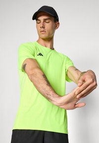Man in een neongroene sportshirt en zwarte pet strekt zijn arm en toont arm tatoeages, tegen een effen lichte achtergrond.