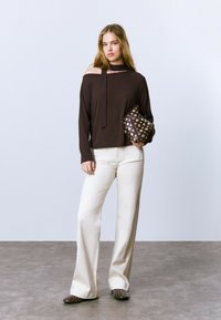Haut marron à manches longues avec une épaule dénudée et un détail noué, associé à un pantalon large blanc et une pochette marron à pois.