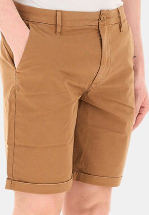 Pantaloncini marroni fino al ginocchio con bottone e zip frontali, tasche laterali, indossati da una persona con una mano in tasca.