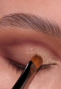 Eine Nahaufnahme eines Auges mit schimmerndem goldenen Lidschatten, der mit einem kleinen Pinsel aufgetragen wurde, wobei sanfte Brauntöne und Mauve in der Lidfalte verblendet sind.