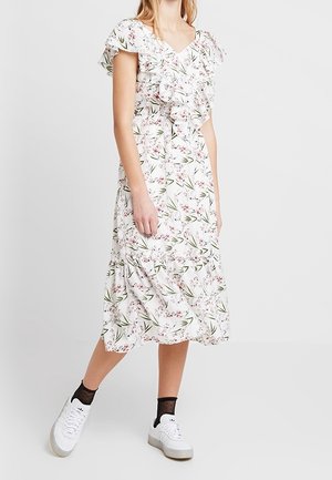 Vrouw draagt een witte midi-jurk met bloemenprint en ruches, witte sneakers en zwarte doorschijnende enkelsokken.