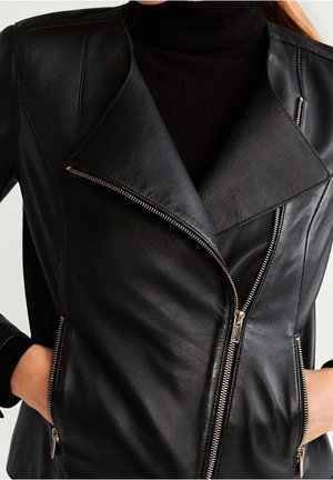 Veste de motard en similicuir noir avec fermeture éclair asymétrique à l'avant et poches latérales zippées, portée sur un pull à col roulé noir.