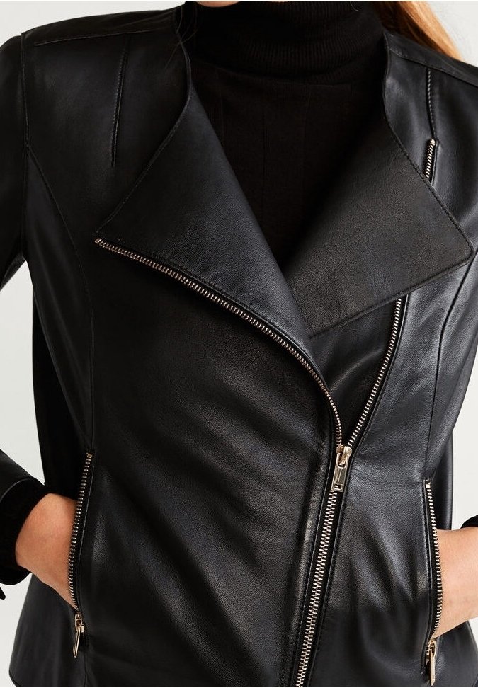 Veste de motard en similicuir noir avec fermeture éclair asymétrique à l'avant et poches latérales zippées, portée sur un pull à col roulé noir.